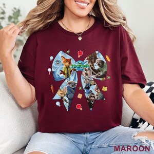 Op de afbeelding: Bordeauxrood T-shirt met een groot strikdesign met personages uit de animatiefilm "Zootopia". De strik is lichtblauw met verschillende personages en het logo van de film. Het woord "MAROON" staat rechtsonder.