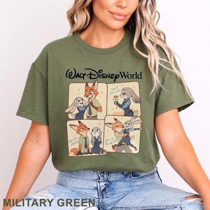 May include: Military green t-shirt featuring a four-panel illustration of Zootopia characters. The top reads "Walt Disney World" in black script. The panels show Nick Wilde and Judy Hopps in various poses. The shirt is made of a soft material.