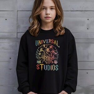 Pode incluir: Camisola preta com um gr&aacute;fico colorido com as palavras "UNIVERSAL STUDIOS" e v&aacute;rios personagens de desenhos animados e um castelo. O gr&aacute;fico est&aacute; num esquema de cores arco-&iacute;ris.
