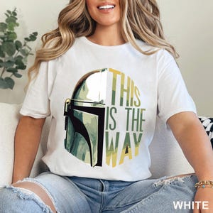 Pu&ograve; includere: Una t-shirt bianca a maniche corte con una stampa grafica di un casco metallico sul lato sinistro e il testo THIS IS THE WAY in lettere maiuscole verdi e gialle sul lato destro, centrato sulla parte anteriore del capo di abbigliamento.
