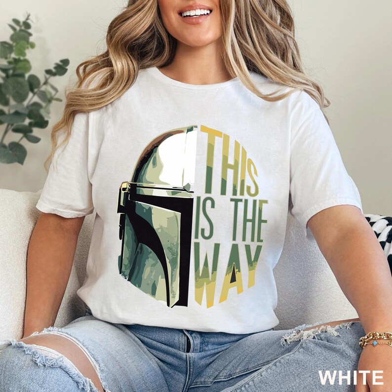 Pu&ograve; includere: Una t-shirt bianca a maniche corte con una stampa grafica di un casco metallico sul lato sinistro e il testo THIS IS THE WAY in lettere maiuscole verdi e gialle sul lato destro, centrato sulla parte anteriore del capo di abbigliamento.