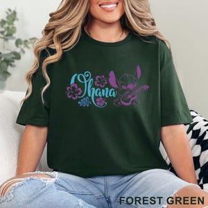 Op de afbeelding: Een bosgroen T-shirt met korte mouwen en een grafische print in lichtblauw en paars. Het ontwerp bevat het woord Ohana in cursief schrift, verschillende hibiscusbloemen en een stripfiguur die op een klein snaarinstrument speelt.