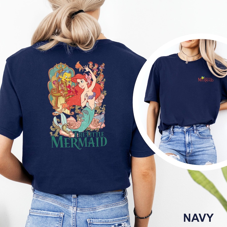 May include: A navy blue short-sleeve t-shirt featuring a large graphic on the back of Ariel and Flounder from The Little Mermaid with floral accents and the text The Little Mermaid. The front chest area displays a small The Little Mermaid logo.