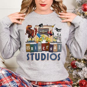 May include: Sport Grey sweatshirt featuring a colorful graphic of Universal Studios characters, including dinosaurs, superheroes, and cartoon figures. The text "Universal Studios" is displayed in a bold, multi-colored font. The sweatshirt is a comfortable, casual style.