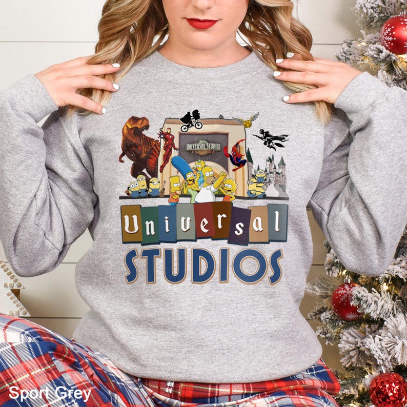 May include: Sport Grey sweatshirt featuring a colorful graphic of Universal Studios characters, including dinosaurs, superheroes, and cartoon figures. The text "Universal Studios" is displayed in a bold, multi-colored font. The sweatshirt is a comfortable, casual style.