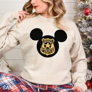 May include: Sand-colored sweatshirt featuring a black Mickey Mouse silhouette with a Zootopia Police badge design. The badge is gold with a star and the words "Zootopia Police". The sweatshirt has long sleeves and a crew neck.