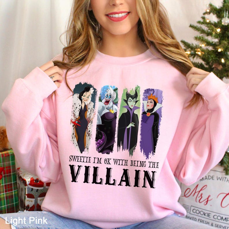 Puede incluir: Sudadera rosa claro con una ilustraci&oacute;n de cuatro villanos de dibujos animados. El texto "SWEETIE I'M OK WITH BEING THE VILLAIN" est&aacute; impreso en negro debajo. La sudadera es de color rosa claro.