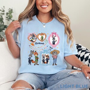 Puede incluir: Una camiseta azul claro con un estampado gr&aacute;fico de Zootopia. El dise&ntilde;o incluye ilustraciones de Judy Hopps y Nick Wilde dentro de marcos en forma de coraz&oacute;n y ornamentados, un icono de zanahoria y el texto Welcome to Zootopia con corazones.