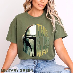 Pu&ograve; includere: Una t-shirt a maniche corte color verde militare con una stampa grafica di un casco metallico sul lato sinistro e il testo THIS IS THE WAY in lettere stampatello gialle e verdi sul lato destro, centrato sul petto del capo.