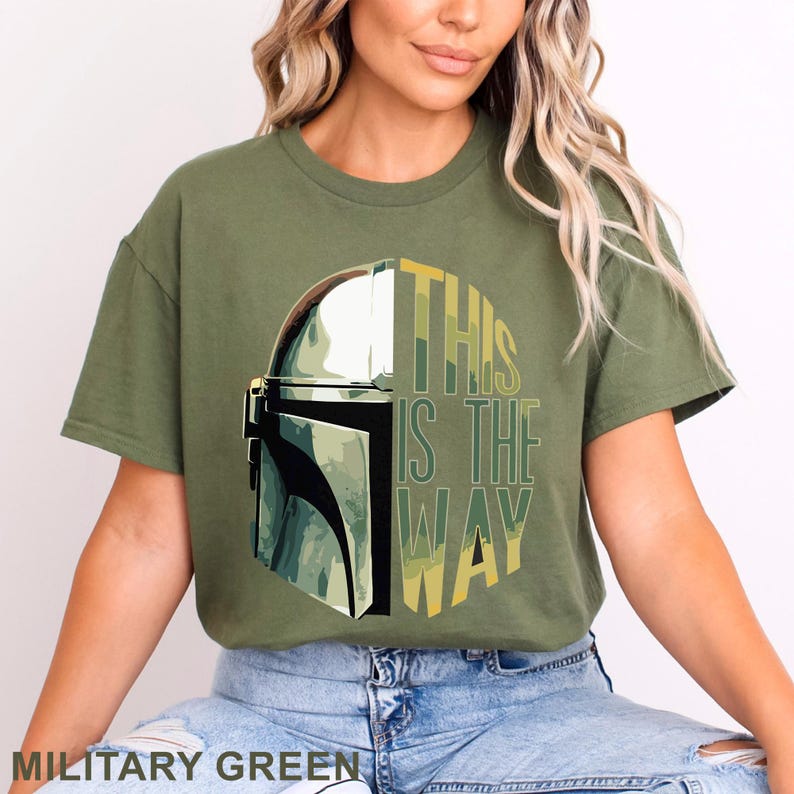 Pu&ograve; includere: Una t-shirt a maniche corte color verde militare con una stampa grafica di un casco metallico sul lato sinistro e il testo THIS IS THE WAY in lettere stampatello gialle e verdi sul lato destro, centrato sul petto del capo.