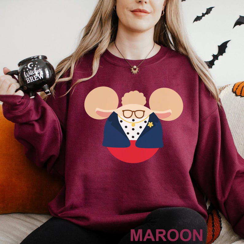 May include: Maroon sweatshirt featuring a cartoon character design with large ears, glasses, and a blazer. The word "MAROON" is printed at the bottom. The person is holding a black mug with the text "Witches Brew".