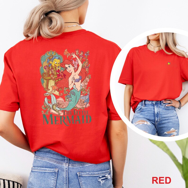 May include: A red short-sleeved t-shirt featuring a large graphic on the back of Ariel from The Little Mermaid with Flounder, surrounded by floral elements and the text The Little Mermaid. The front features a small embroidered character icon.