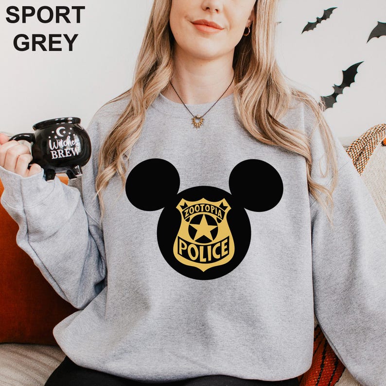 May include: Sport grey sweatshirt featuring a black Mickey Mouse silhouette with a Zootopia Police badge design in gold and black. The sweatshirt has a crew neck and long sleeves. A black mug with the text "Witches Brew" is held in the left hand.