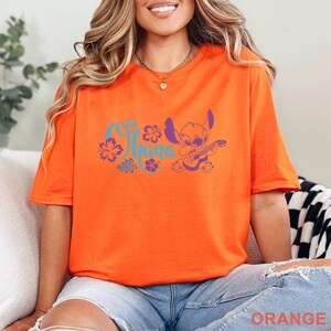 Op de afbeelding: Een feloranje T-shirt met korte mouwen en een paarse afbeelding van het personage Stitch die op een ukelele speelt. Het ontwerp bevat het woord Ohana in een gestileerd cursief lettertype, omringd door paarse hibiscusbloemen en bladeren.