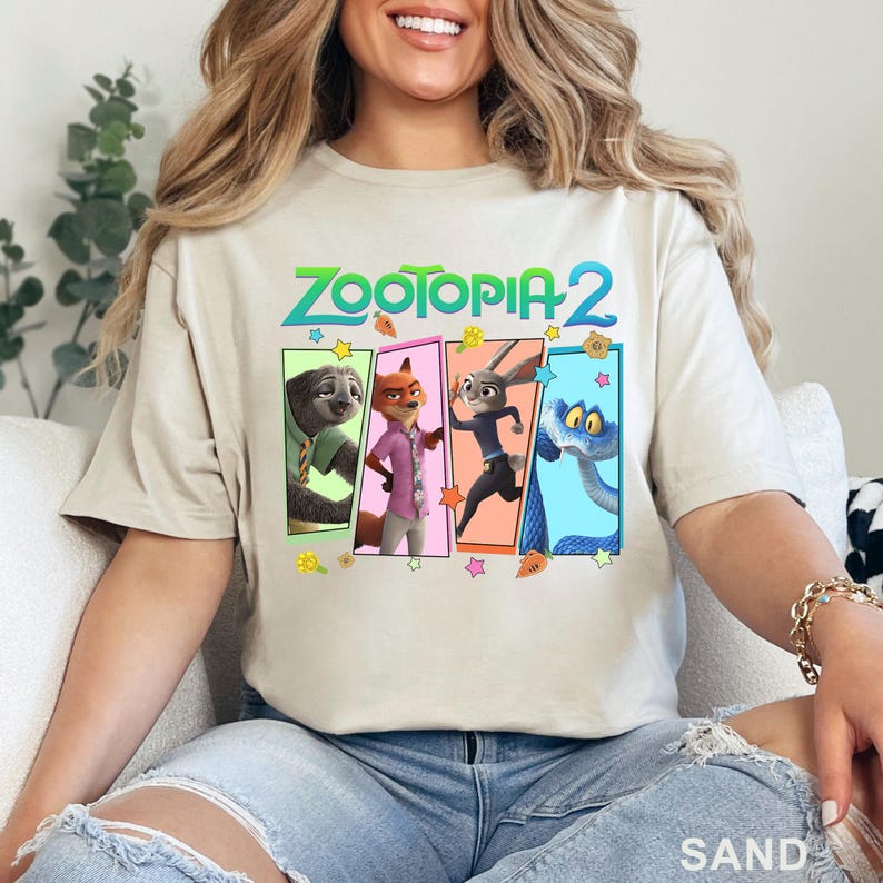 May include: Sand-coloured t-shirt with the text "ZOOTOPIA 2" in green and blue. Below the text are four character illustrations from the film, each in a different coloured rectangle. The shirt is a crew neck style.