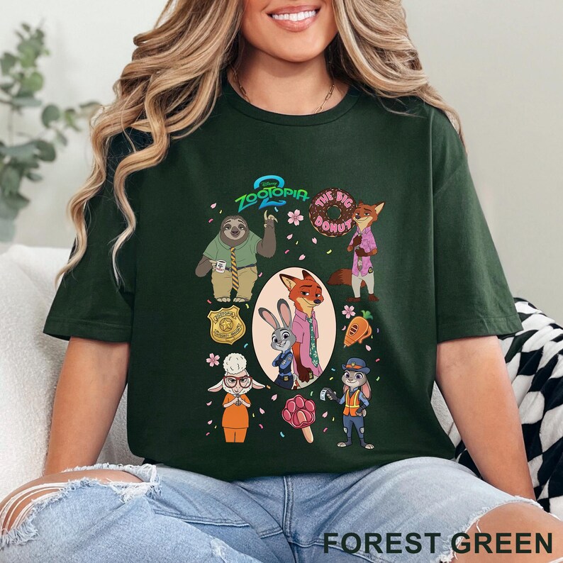 K&ouml;nnte beinhalten: Ein dunkelwaldgr&uuml;nes T-Shirt mit einem Zootopia 2 Grafikdruck. Das Design zeigt Charaktere wie Nick Wilde, Judy Hopps und Flash sowie einen Donut, ein Abzeichen und ein pfotenf&ouml;rmiges Eis am Stiel, umgeben von kleinen rosa Bl&uuml;tenbl&auml;ttern.