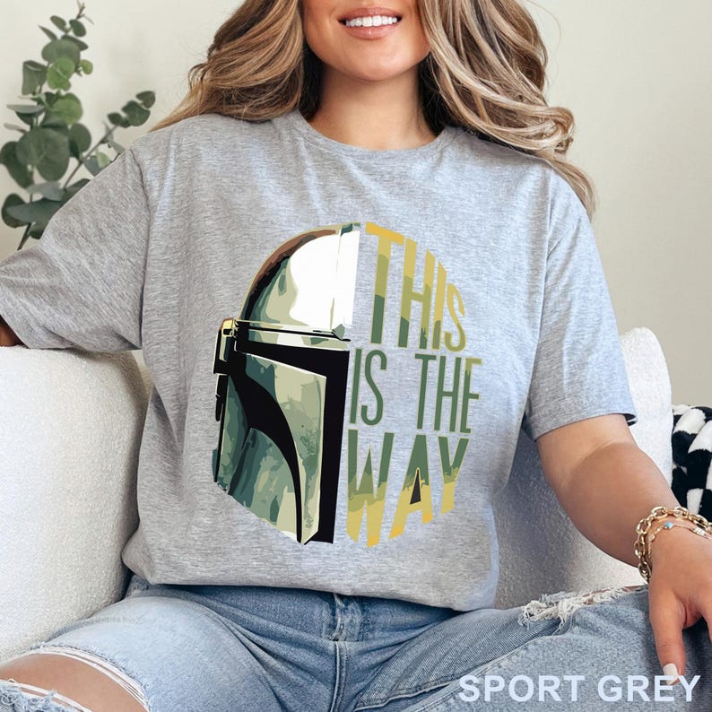 Pu&ograve; includere: Una t-shirt a maniche corte color grigio sportivo con la grafica di un elmo mandaloriano sul lato sinistro e il testo "THIS IS THE WAY" in lettere maiuscole gialle e verdi sul lato destro, su uno sfondo circolare con motivo mimetico.