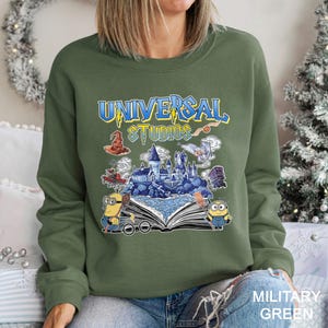 Op de afbeelding: Olijfgroene sweatshirt met een grafisch ontwerp van het Universal Studios-logo met illustraties van een kasteel, Minions en andere themaparkelementen. De tekst "UNIVERSAL STUDIOS" is in blauw en geel. Het sweatshirt is gelabeld "MILITARY GREEN".
