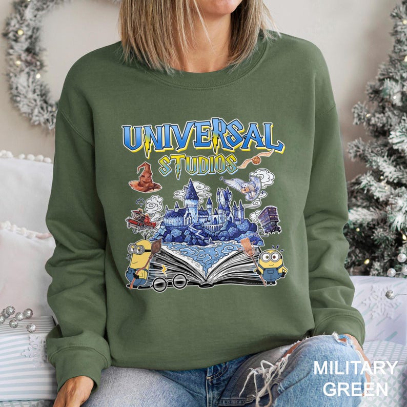 Op de afbeelding: Olijfgroene sweatshirt met een grafisch ontwerp van het Universal Studios-logo met illustraties van een kasteel, Minions en andere themaparkelementen. De tekst "UNIVERSAL STUDIOS" is in blauw en geel. Het sweatshirt is gelabeld "MILITARY GREEN".