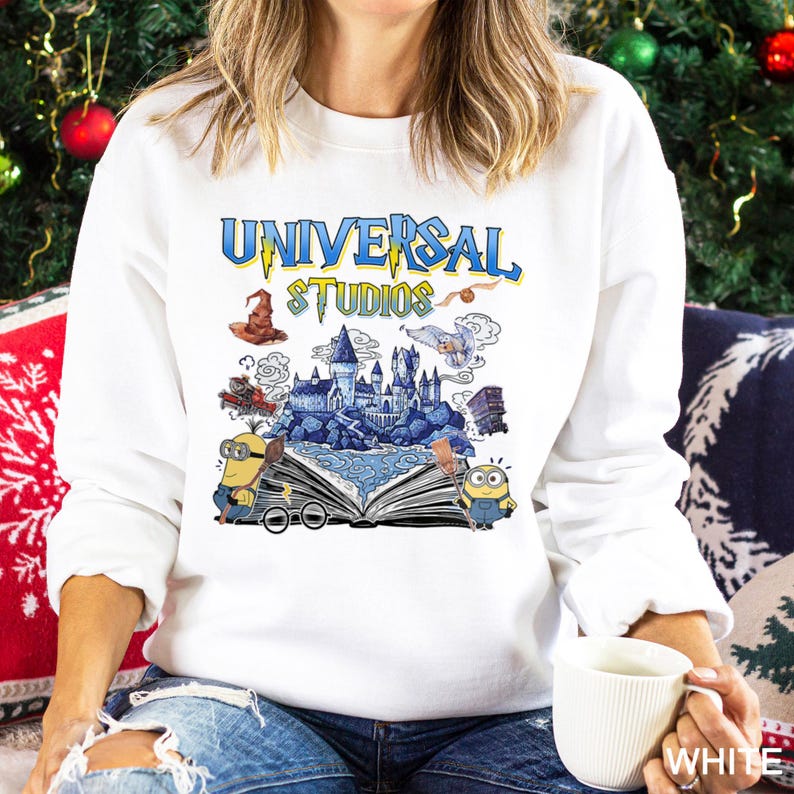Op de afbeelding: Witte sweatshirt met een afbeelding met "UNIVERSAL STUDIOS" in blauwe en gele letters. Het ontwerp bevat illustraties van een kasteel, Minions en andere thematische elementen.