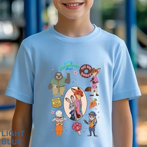 K&ouml;nnte beinhalten: Ein hellblaues Kurzarm-T-Shirt mit einem Zootopia 2 Grafikdruck. Das Design zeigt Charaktere wie Judy Hopps, Nick Wilde und Flash sowie einen Donut, ein Abzeichen, eine Karotte und ein pfotenf&ouml;rmiges Eis am Stiel, umgeben von rosa Bl&uuml;ten.