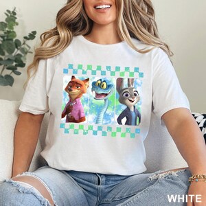 May include: White t-shirt featuring a colorful graphic of three cartoon characters: a fox in a pink shirt, a blue snake, and a rabbit in a blue jacket. The graphic is framed by a blue and green checkered pattern. The word "WHITE" is printed at the bottom.