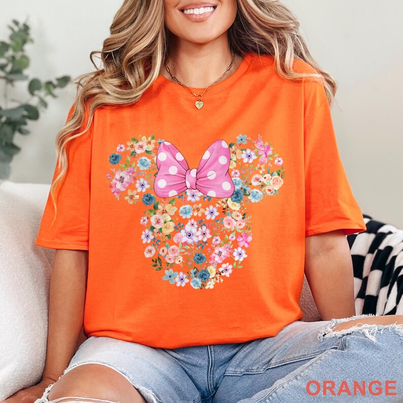 May include: Bright orange t-shirt featuring a floral design in the shape of a mouse head with a pink polka dot bow. The design is made up of various colorful flowers. The word "ORANGE" is printed at the bottom of the shirt.