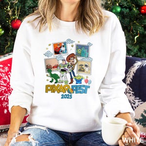 Może przedstawiać: Biała bluza z kolorową grafiką postaci z film&oacute;w Pixar i napisem "PIXAR FEST 2025". Projekt zawiera obrazy Woody'ego, Buzza Astrala i innych postaci z film&oacute;w Pixar. Bluza jest w stylu casual.