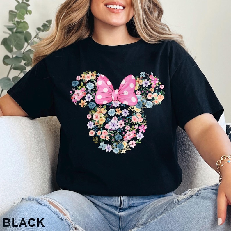 May include: Black t-shirt featuring a floral Minnie Mouse design. The design includes a pink polka-dot bow and a floral arrangement in the shape of Minnie's head. The word "BLACK" is printed at the bottom.