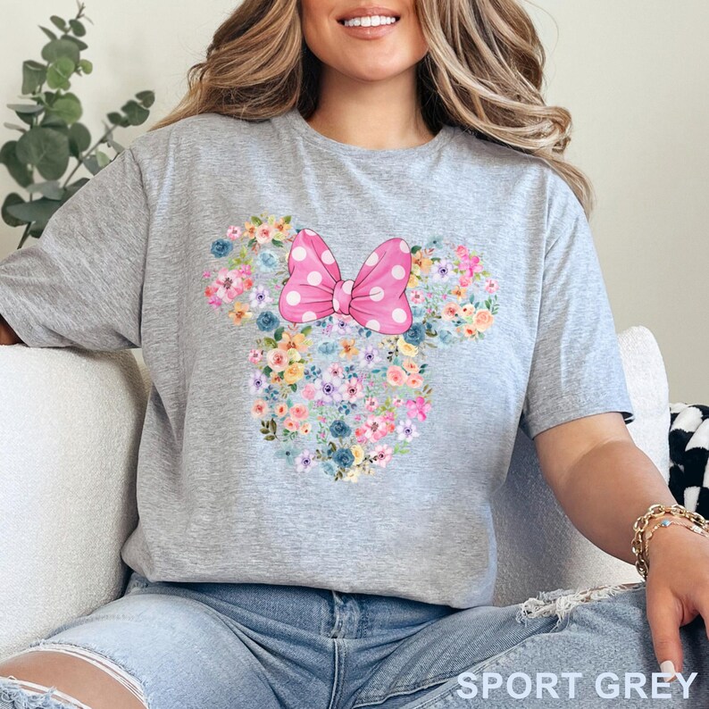 May include: Sport grey t-shirt featuring a floral Minnie Mouse design. The design includes a pink bow with white polka dots and a floral arrangement in the shape of Minnie's head. The shirt is made of a soft material.