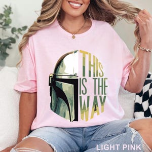 Pu&ograve; includere: Una t-shirt rosa chiaro con una grafica di un casco metallico sul lato sinistro e il testo "THIS IS THE WAY" in lettere stampatello verdi e gialle sul lato destro. Il design &egrave; centrato sul petto del capo a maniche corte.