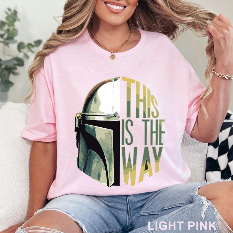 Pu&ograve; includere: Una t-shirt rosa chiaro con una grafica di un casco metallico sul lato sinistro e il testo "THIS IS THE WAY" in lettere stampatello verdi e gialle sul lato destro. Il design &egrave; centrato sul petto del capo a maniche corte.