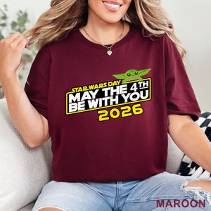 May include: Maroon t-shirt with the text "STAR WARS DAY MAY THE 4TH BE WITH YOU 2026" in white and yellow, with a green cartoon character. The shirt is a crew neck.