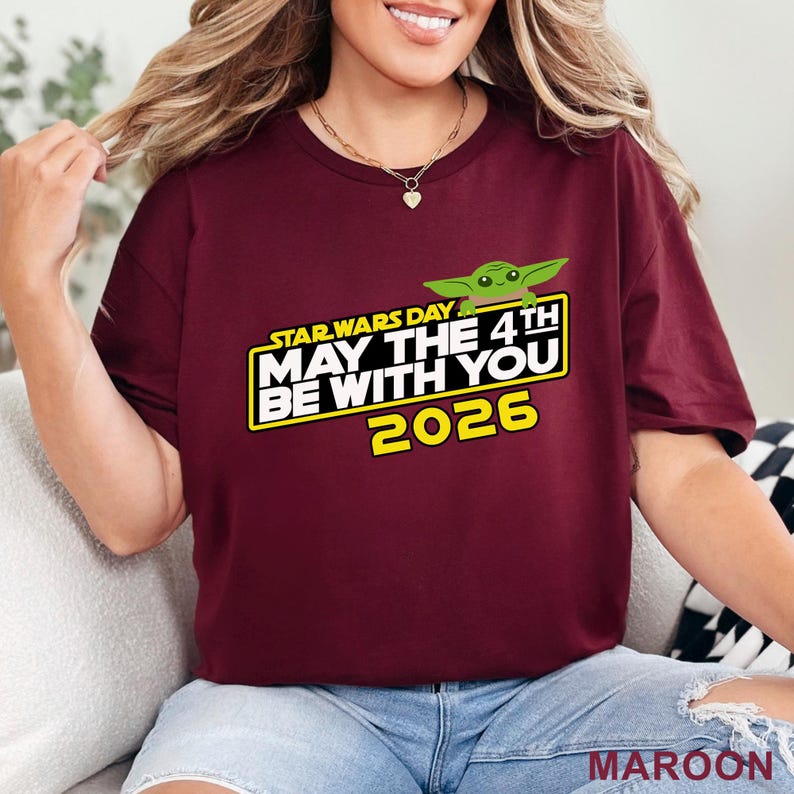 May include: Maroon t-shirt with the text "STAR WARS DAY MAY THE 4TH BE WITH YOU 2026" in white and yellow, with a green cartoon character. The shirt is a crew neck.