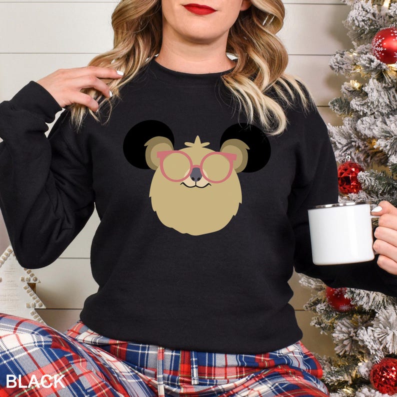 May include: Black crewneck sweatshirt featuring a cartoon character with large black ears, tan fur, and pink glasses. The word "BLACK" is printed at the bottom. The sweatshirt is part of a clothing collection.