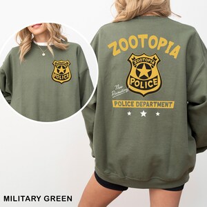 May include: Military green sweatshirt featuring the text "ZOOTOPIA" and a police badge graphic on the back and front. The badge has a star in the center and the word "POLICE". The sweatshirt also has the text "POLICE DEPARTMENT" and "Now Recruiting".