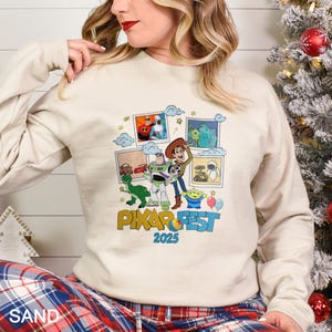Może przedstawiać: Beżowa bluza z kolorową grafiką postaci z film&oacute;w Pixar. Projekt zawiera wizerunki Buzza Astrala, Chudego i Sully'ego, z napisem "PIXAR FEST 2025" w zabawnej czcionce. Bluza ma długie rękawy.