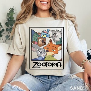 May include: Sand-coloured t-shirt with a framed graphic of characters from the animated film "Zootopia." The graphic includes a fox, a rabbit, and other animal characters. The word "Zootopia" is printed in a bold, black font below the characters.