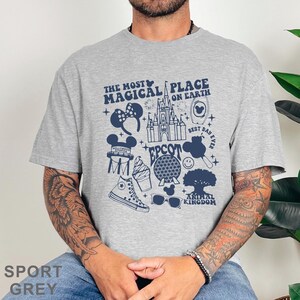 May include: Sport grey t-shirt featuring a navy blue graphic design with the text "The Most Magical Place on Earth." The design includes icons of a castle, Mickey Mouse ears, a sneaker, and other theme park elements.