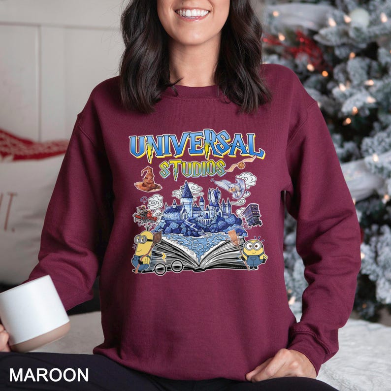 Op de afbeelding: Bordeaux sweatshirt met een kleurrijke afbeelding van het Universal Studios-logo, een kasteel en Minions. Het ontwerp is geplaatst tegen een open boek. De tekst "UNIVERSAL STUDIOS" is in blauw en geel.