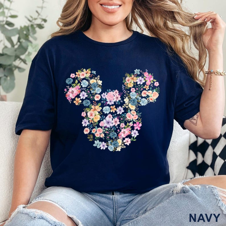 May include: Navy blue t-shirt featuring a floral Mickey Mouse design. The design is composed of colorful flowers in shades of pink, blue, yellow, and white. The shirt has short sleeves and a relaxed fit. The word "NAVY" is printed at the bottom.