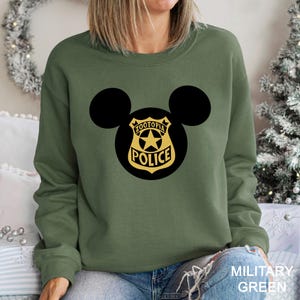 May include: Military green sweatshirt featuring a black Mickey Mouse silhouette with a Zootopia Police badge design in black and gold. The text "MILITARY GREEN" is visible at the bottom right.