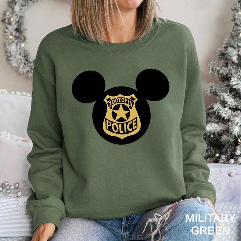 May include: Military green sweatshirt featuring a black Mickey Mouse silhouette with a Zootopia Police badge design in black and gold. The text "MILITARY GREEN" is visible at the bottom right.