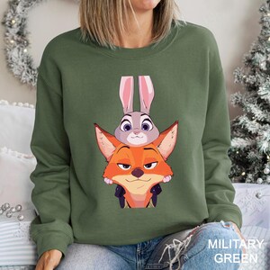 May include: Military green sweatshirt featuring a graphic of the Zootopia characters Judy Hopps and Nick Wilde. The design shows Judy Hopps, a gray rabbit, atop Nick Wilde, an orange fox. The sweatshirt is a crew neck style.