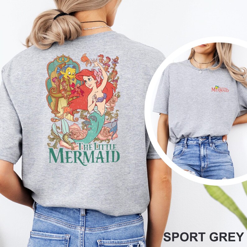 May include: A sport grey t-shirt featuring a large graphic on the back of Ariel from The Little Mermaid surrounded by floral elements and sea creatures. The front chest area displays a small logo with the text The Little Mermaid in colorful font.