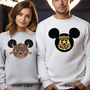 May include: Two sweatshirts, one sport grey and one white, each featuring a cartoon design. The sport grey sweatshirt has a brown character with orange flowers and black ears. The white sweatshirt displays a black Mickey Mouse head with a Zootopia Police badge.