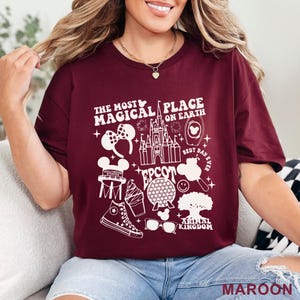 May include: Maroon t-shirt with a white graphic design featuring Disney icons and the text "THE MOST MAGICAL PLACE ON EARTH". The design includes a castle, Mickey Mouse ears, and other park-related images. The word "MAROON" is at the bottom.