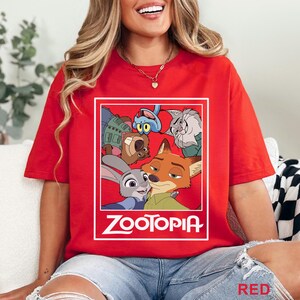 May include: Red t-shirt with a graphic of characters from the animated film "Zootopia" within a white rectangular frame. The word "Zootopia" is printed in white below the graphic. The shirt is a plain red colour.
