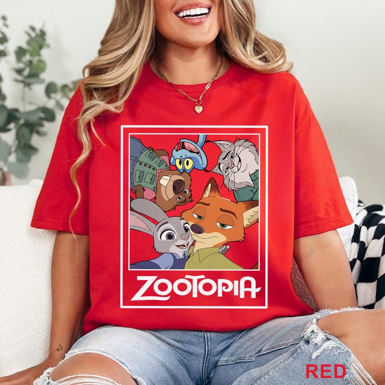 May include: Red t-shirt with a graphic of characters from the animated film "Zootopia" within a white rectangular frame. The word "Zootopia" is printed in white below the graphic. The shirt is a plain red colour.