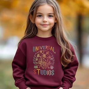 Pode incluir: Um moletom bord&ocirc; com o texto "UNIVERSAL STUDIOS" em um design colorido e vintage. O gr&aacute;fico inclui ilustra&ccedil;&otilde;es de personagens e atra&ccedil;&otilde;es. O moletom &eacute; usado por uma pessoa jovem com cabelos longos e castanhos.
